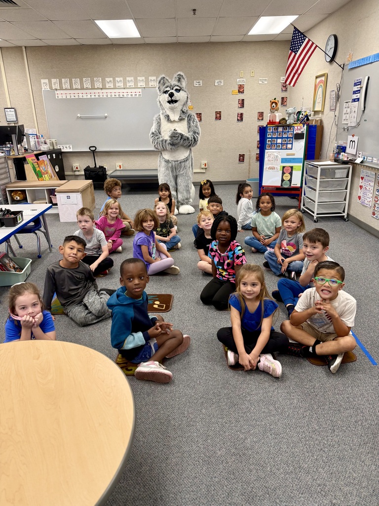Kids in a classroom, seated around a mascot on the floor. The room has a whiteboard, tables, and shelves.