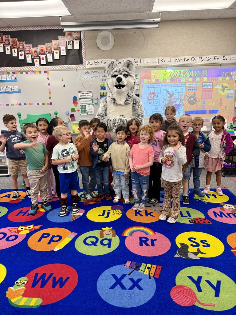 A group of children in a classroom with a mascot standing in the middle. Alphabet letters and pictures on the floor.