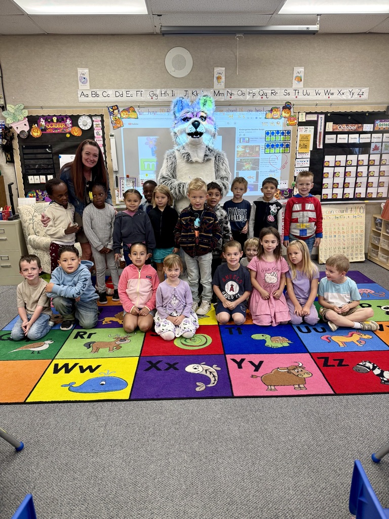 Kindergarteners sit on a colorful alphabet mat with a mascot, in a classroom with a teacher and bulletin boards.