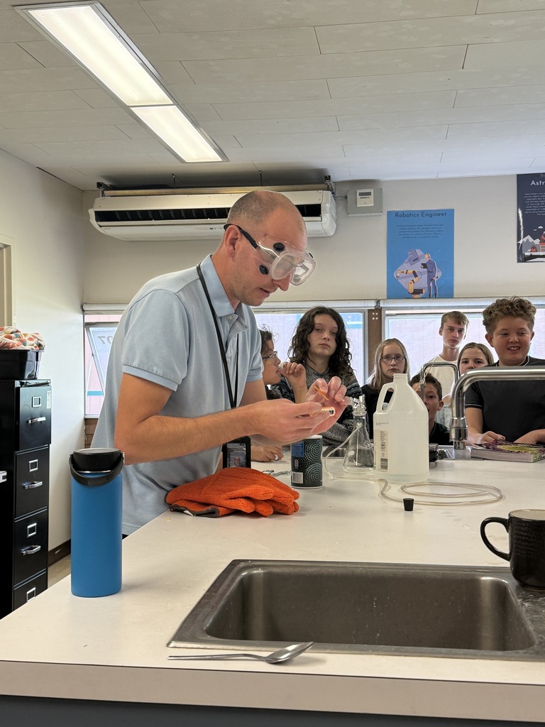 A person stands at a counter in a classroom, holding objects and wearing glasses. A sink is nearby.