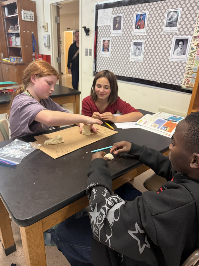 Three students work on a project together, seated around a table. One uses a clay tool.