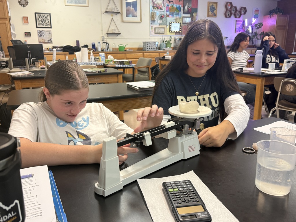 Two students use a scale to measure objects. One girl smiles at the camera.