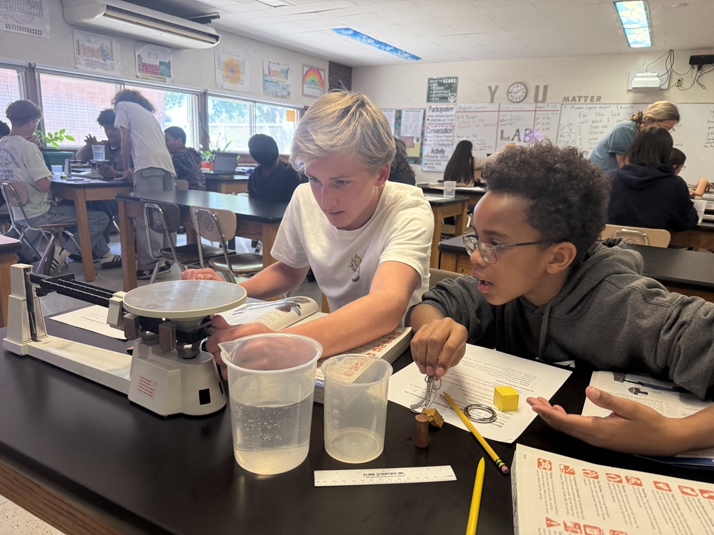 Two students work together in a classroom. They examine a scale, liquids in cups, and a paper.