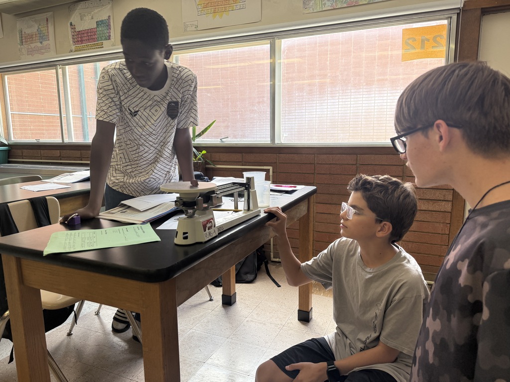 Three boys in a classroom. One boy uses a scale to weigh an object while another observes.