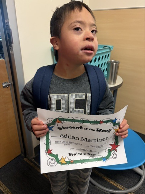 Boy with a blue backpack holding a certificate with the name Adrian Martinez. He is standing in front of a door.