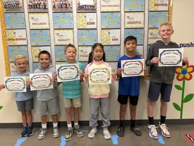 Five young children stand holding certificates. The wall behind them is decorated with pictures and flowers.