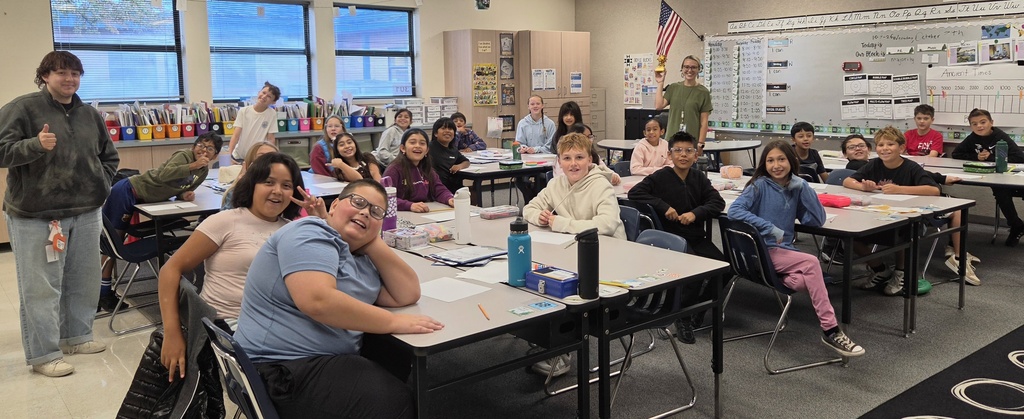 A classroom with several students seated at desks, one person standing, and a whiteboard in the background.