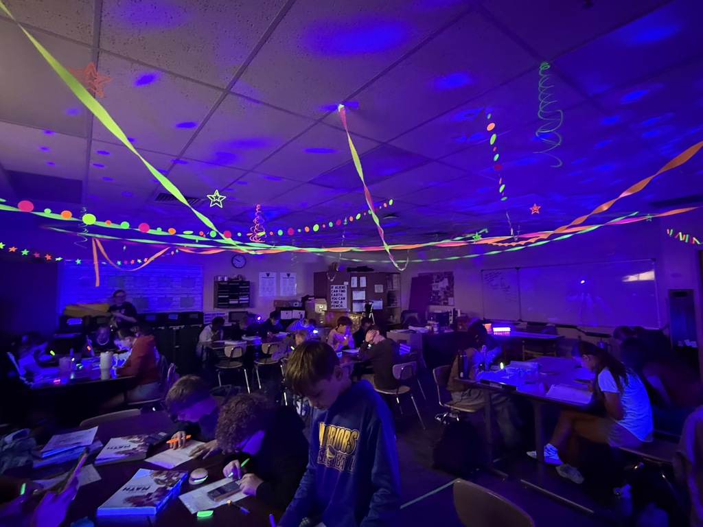 students at tables in a room with tables, chairs, and neon-colored streamers on the ceiling.