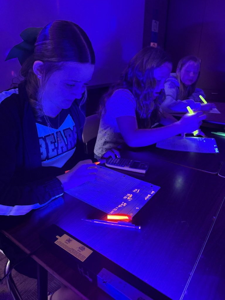 Three individuals seated at a table, using a UV light and pen to draw on paper.
