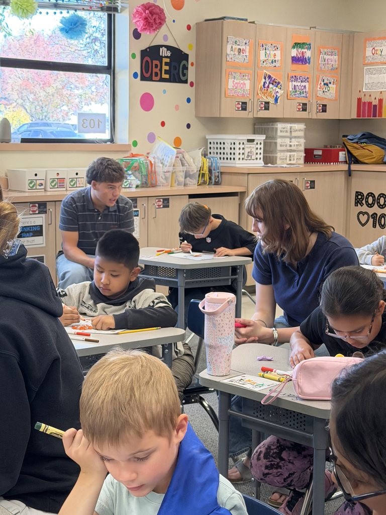 A classroom with students sitting at tables. One student is coloring. A woman is assisting a boy.