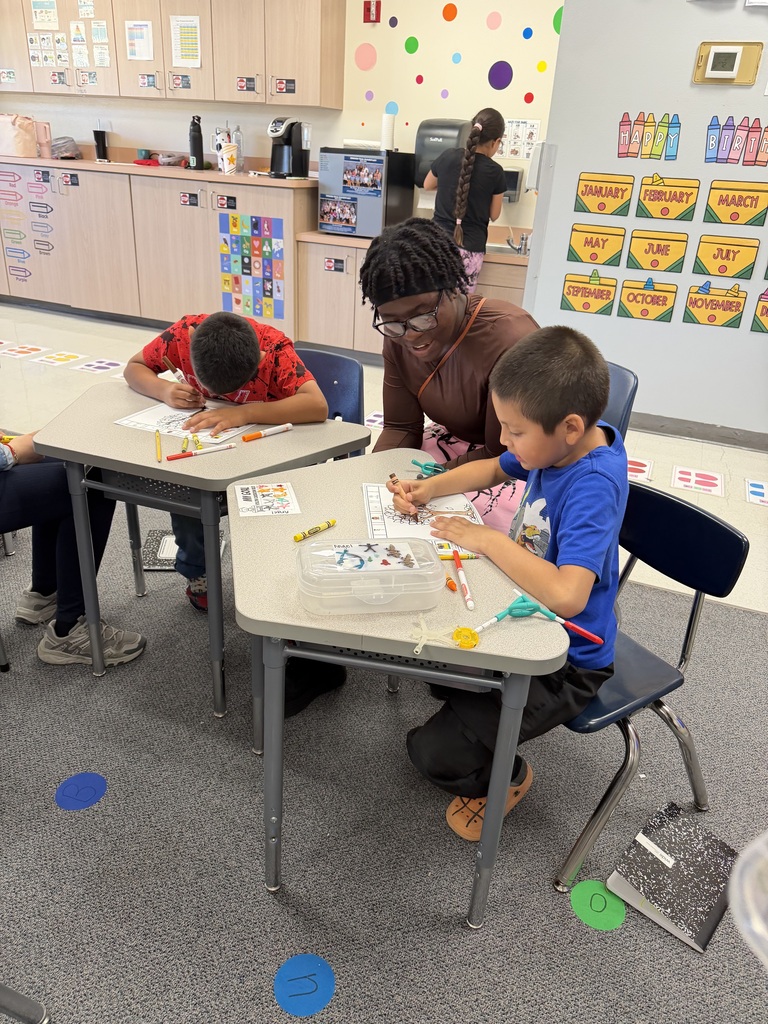 Kids at desks with a woman, possibly a teacher, in a classroom with a colorful wall.