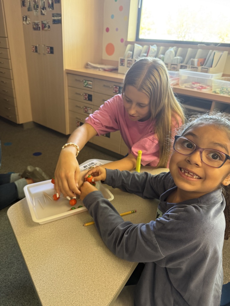 Two children seated at a table, one in glasses, both smiling and working with small objects.