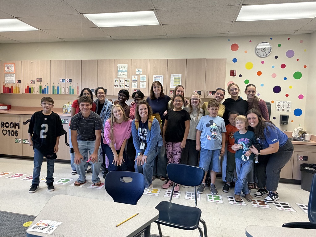 A group of students and adults stand together in a classroom, smiling for a photo. Brightly colored dots adorn the walls.