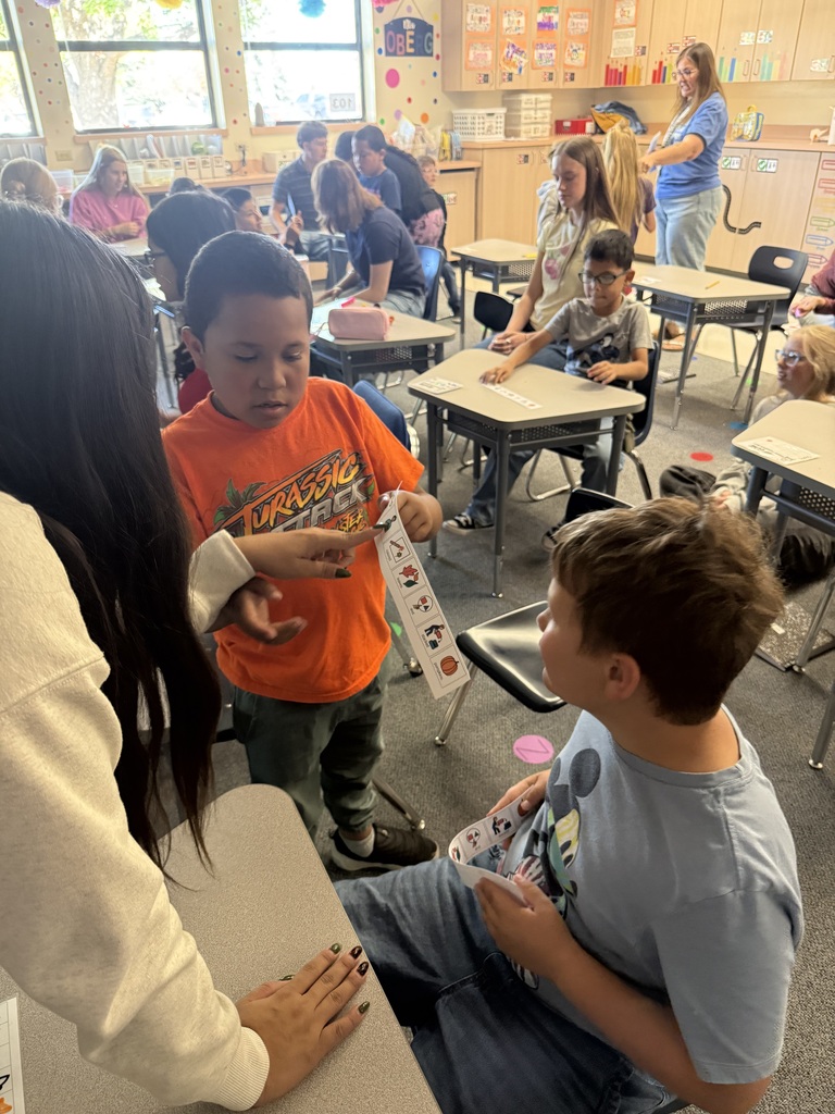 Two children interact with a teacher in a classroom setting, one holding a card with text.