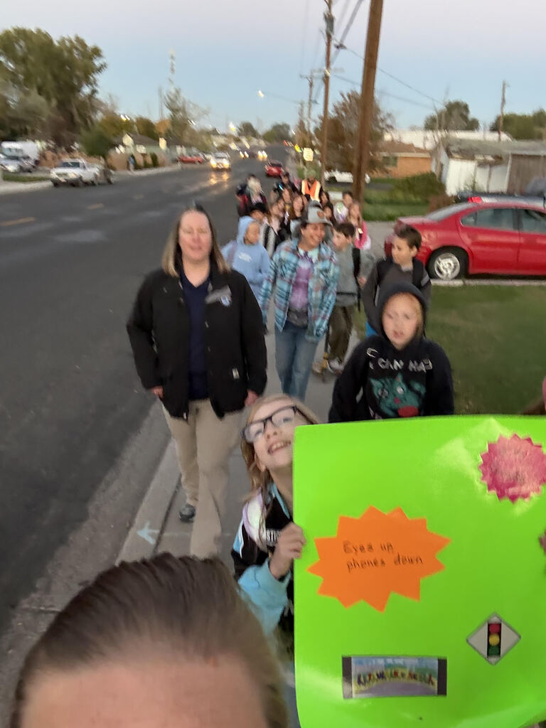 A group of people, including children, walk on a sidewalk, holding a green sign. Behind them, a red car and streetlights are visible.