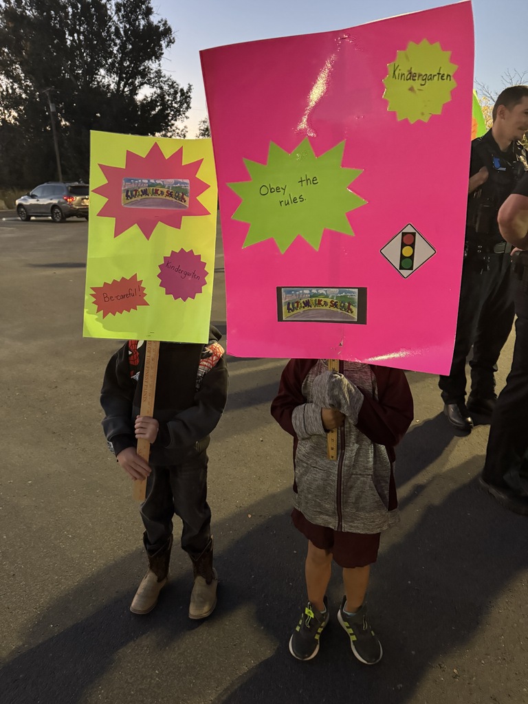 Two children hold pink signs, one featuring a stoplight, in an outdoor area. Behind them, a police officer stands with a vehicle in the distance.