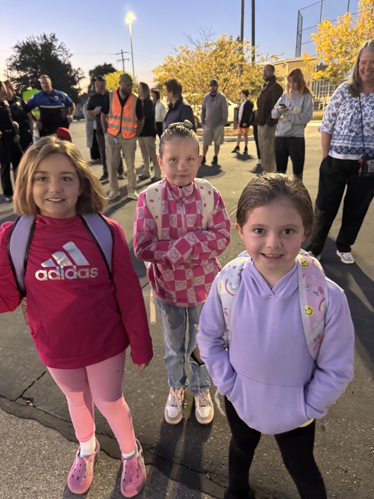 Three girls stand on a road, one with a backpack and another wearing Adidas. People in the background are standing and talking.