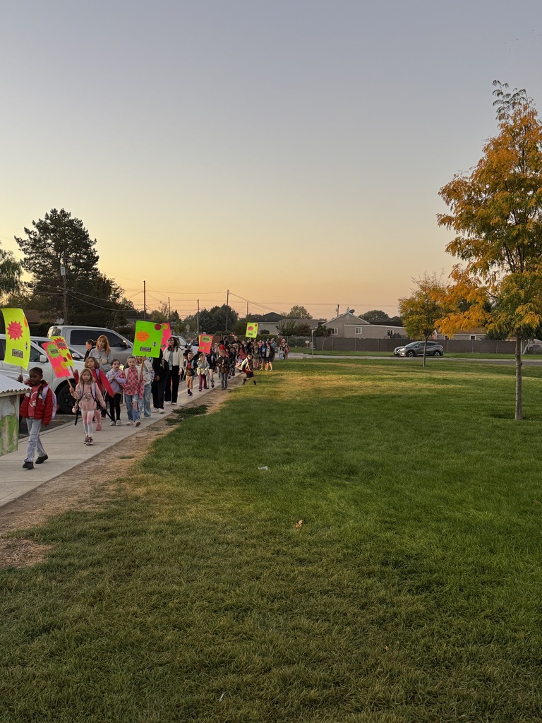 A group of people holding signs walking on a sidewalk near a grassy area. Trees and a sunset are visible in the background.