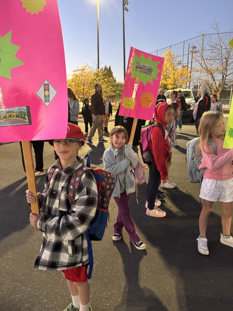 Children hold up pink signs in a parking lot, with trees in background. Some wear backpacks.