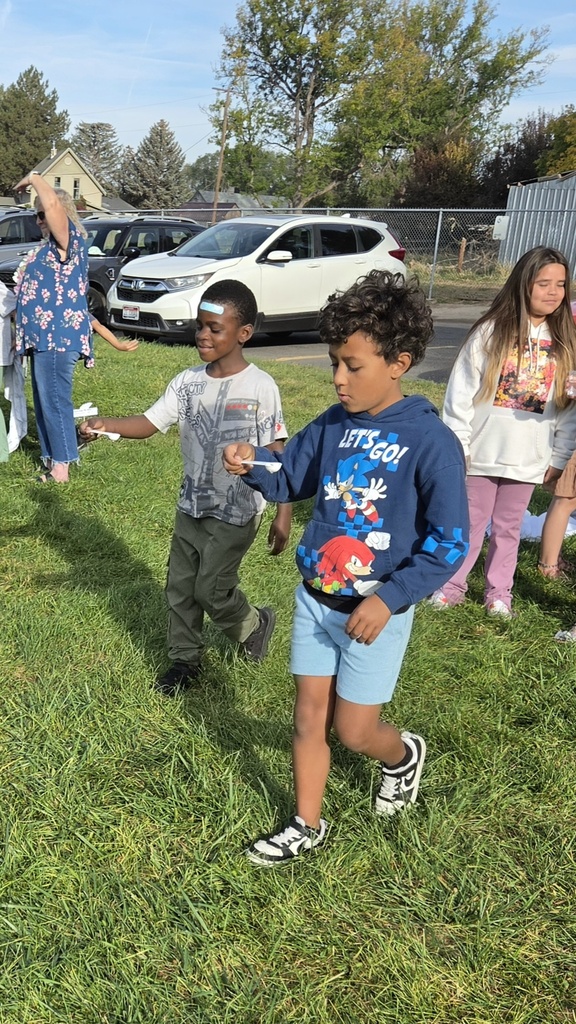 Children play in a grassy area near parked vehicles. One child holds a spoon. A woman in floral clothing stands near a fence.