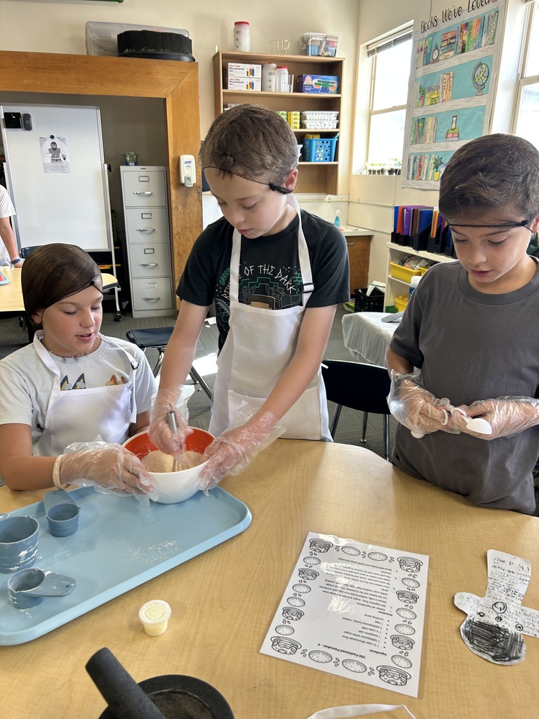 Three students, two wearing aprons and gloves, are mixing a bowl of food on a table.