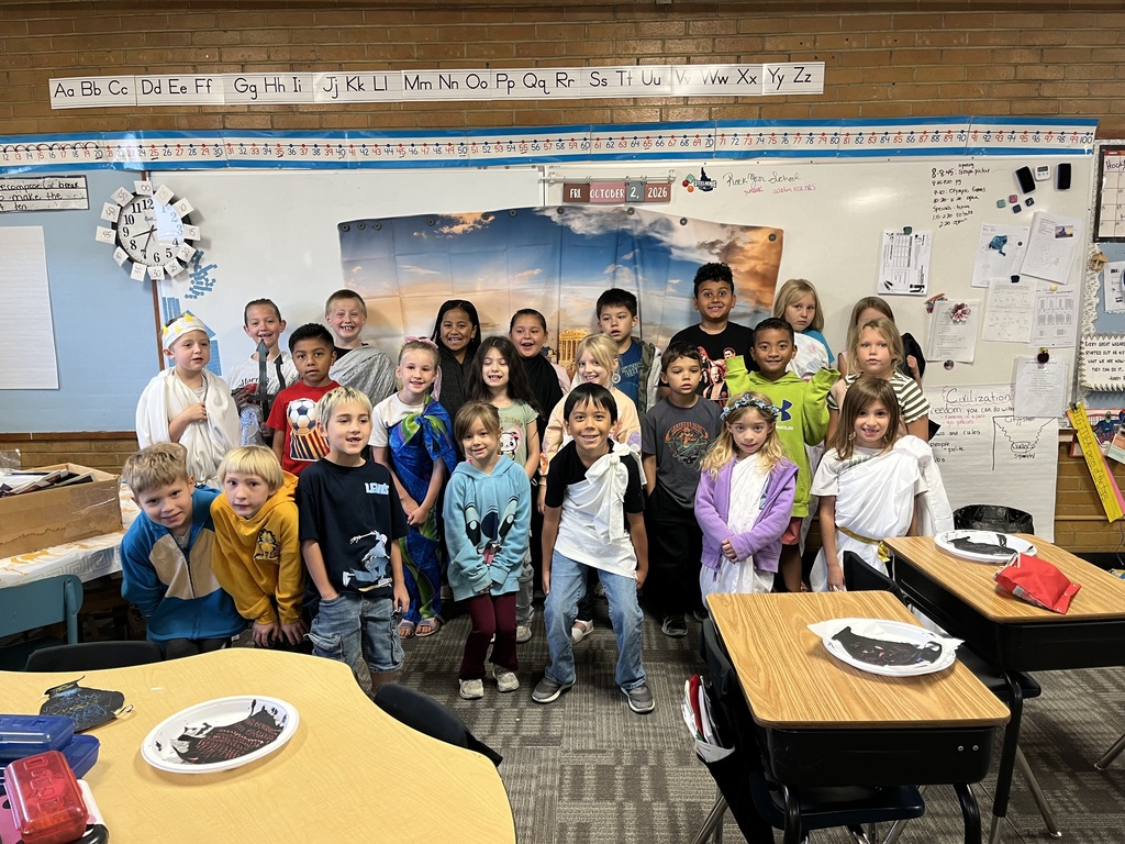 Classroom with students in various outfits, some wearing togas. Desks and chairs are in front.