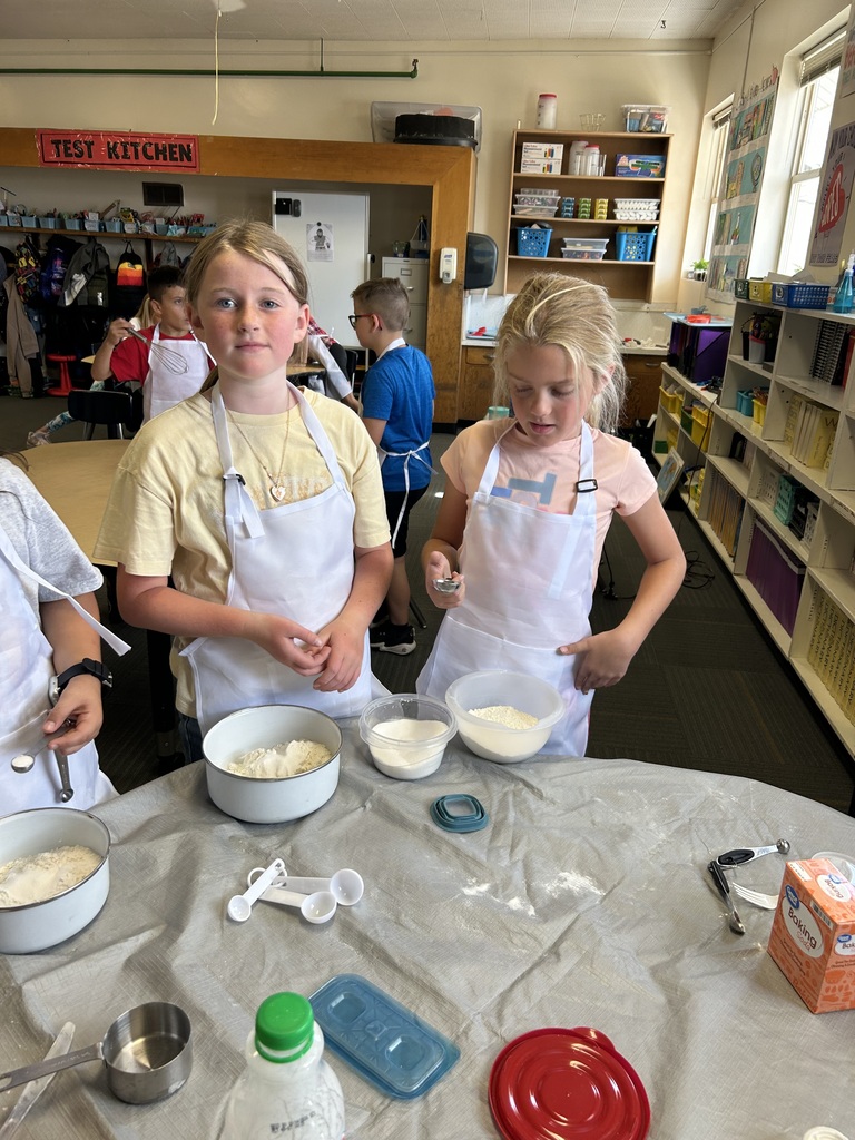 Kids in aprons mix ingredients in bowls at a table, with shelves and other children in the background.