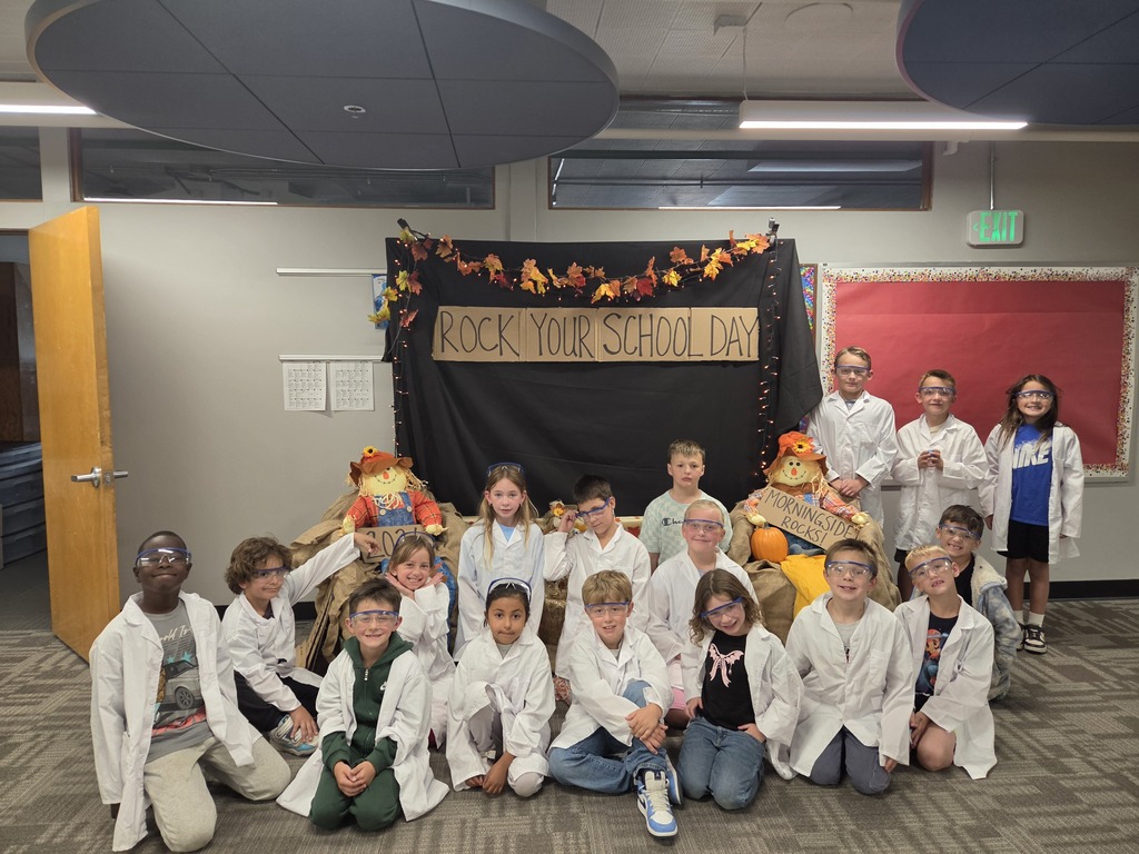 A classroom with children in lab coats sitting and kneeling in rows in front of a banner.