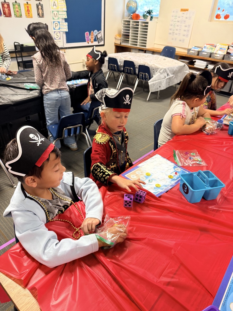 Children at a table in a classroom. Some wear pirate hats and costumes. One holds dice.