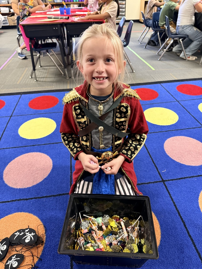 A child in a pirate costume holds a box of candy. They sit on a colorful carpet in a classroom.