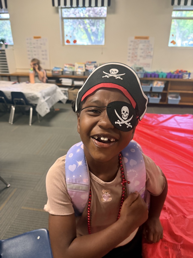 A child wearing a pirate hat and eye patch smiles in a room with tables, chairs, and shelves.