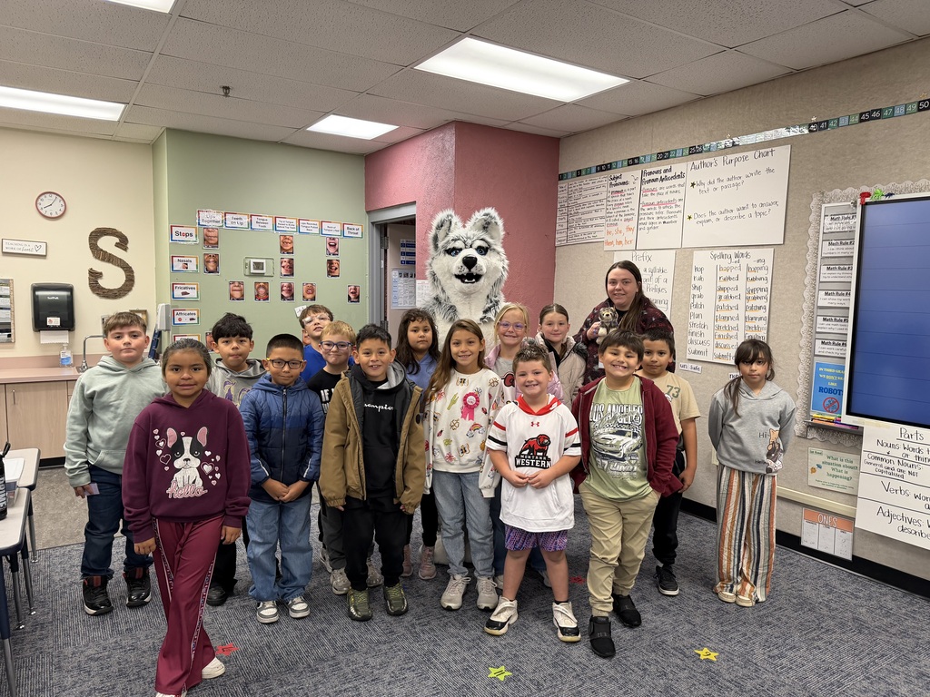 Students stand in a room with a mascot. A woman holds a mascot. Bulletin boards and a large monitor are in the background.