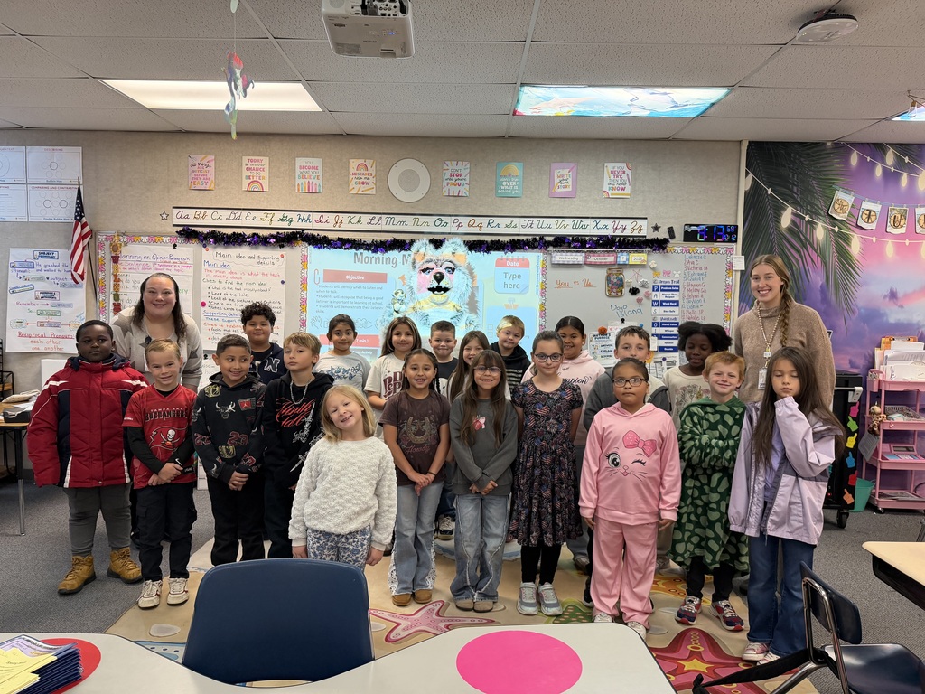 Classroom filled with children, two adults, and desks. Children wearing pajamas. Wall displays and projector screen at the front.