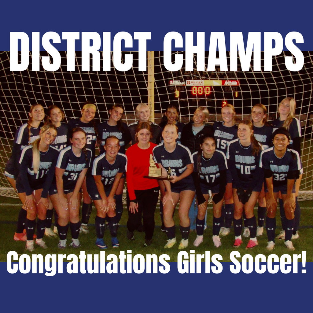 A team of girls' soccer players in blue uniforms stands behind a goal net with a trophy.
