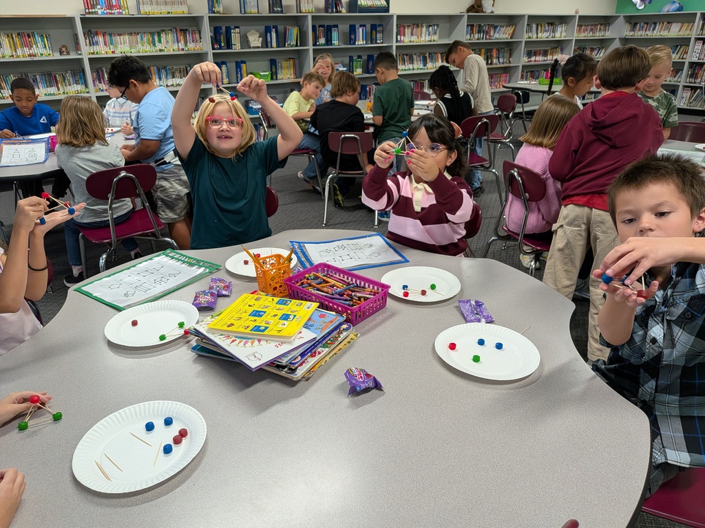 Several children are seated at a round table in a library, engaging in a board game. Plates and toys are on the table.