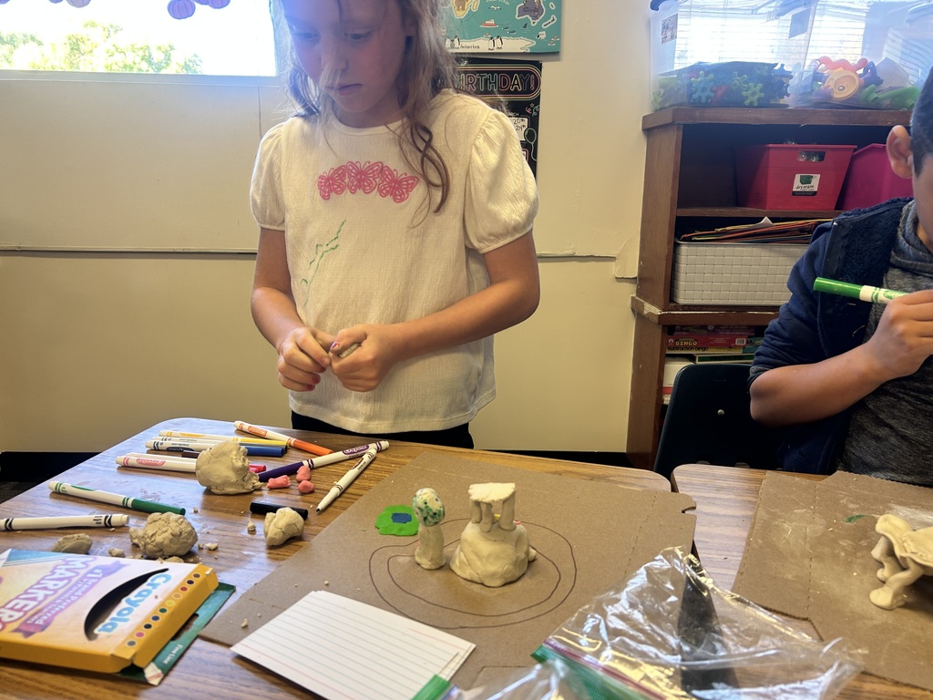 Two children work on a clay sculpture in a classroom. The table holds clay, pens, and a book.