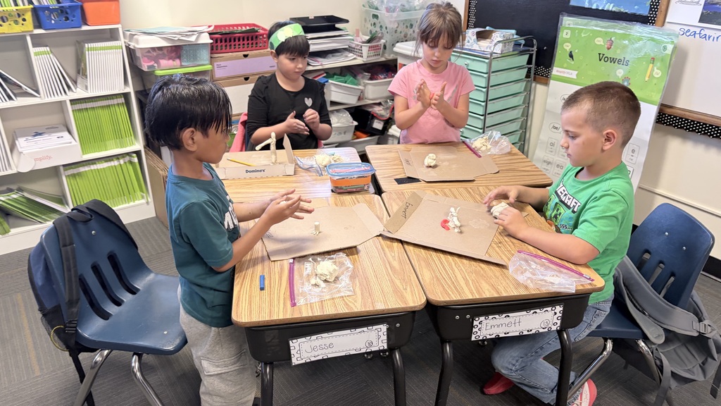 Four children in a classroom sit at desks, engaged in an activity involving paper and small items.