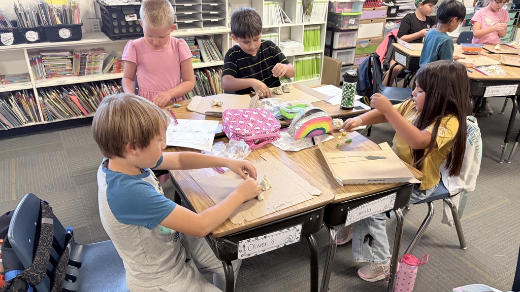 Children sit around tables, engaged in an art activity. Bookshelves and chairs are in the background.