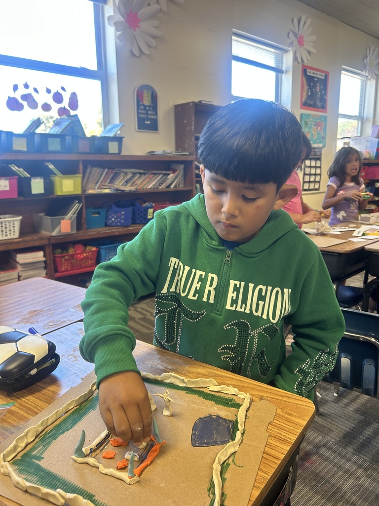 A young child in a green hoodie works on a table, surrounded by classroom furniture.