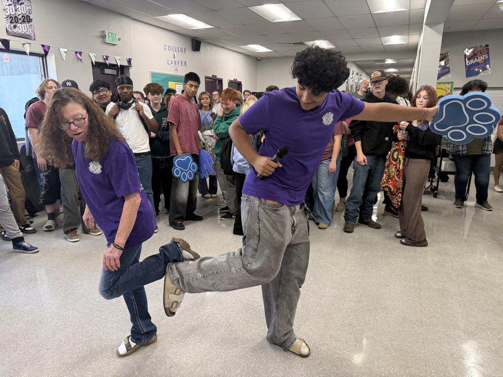 A pair of individuals wearing matching purple shirts and blue gloves perform dance moves in a hallway.