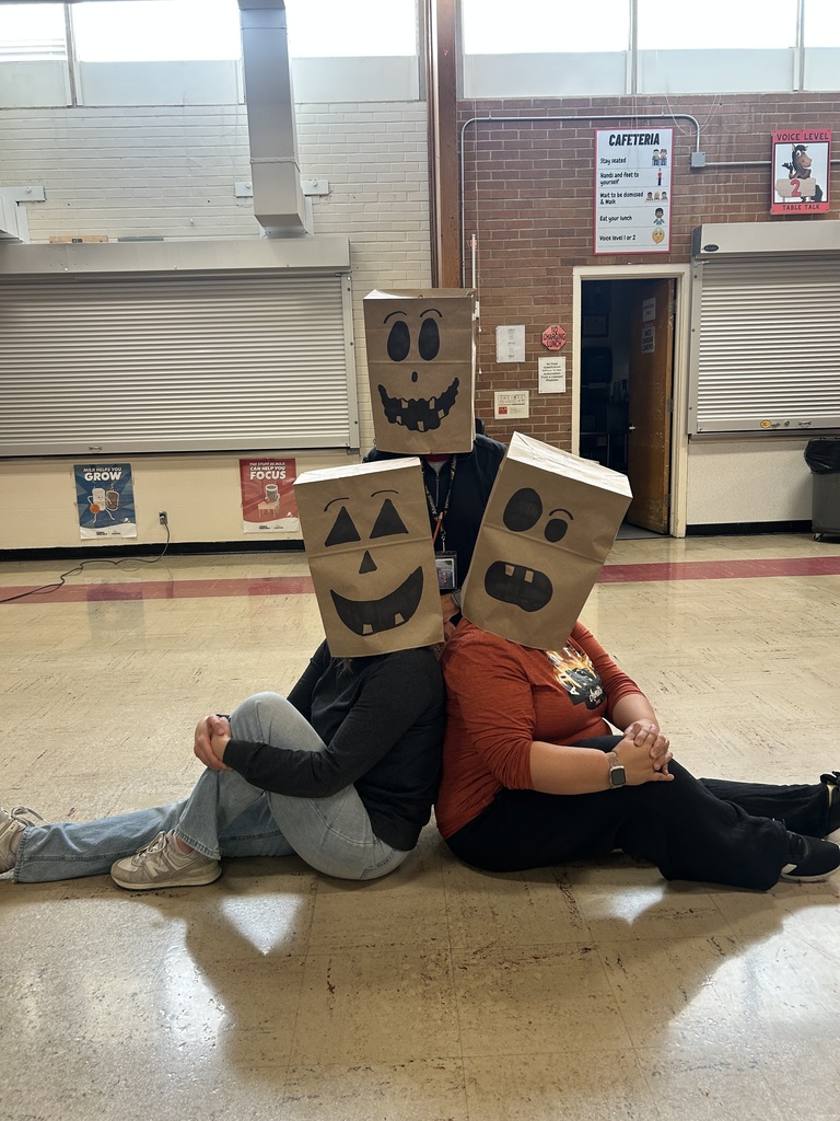 Two people wearing paper bags with faces drawn on them sit on the floor in a room.