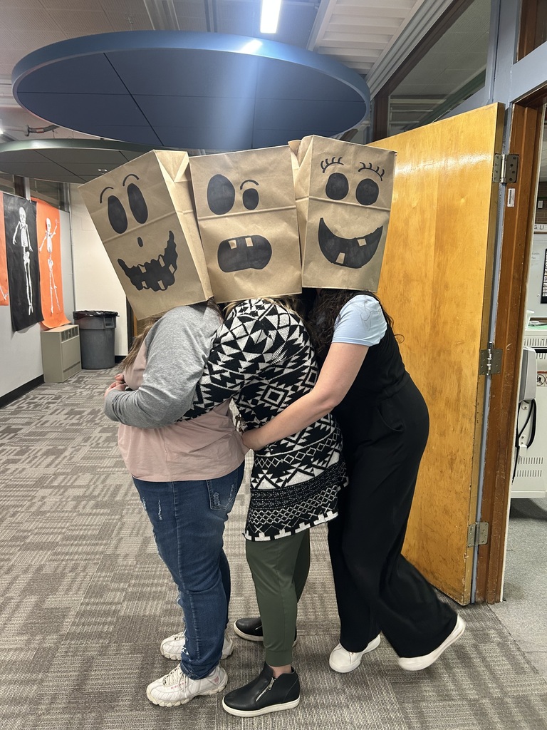 Three people wearing paper bags with drawn faces on them are hugging each other in a hallway.