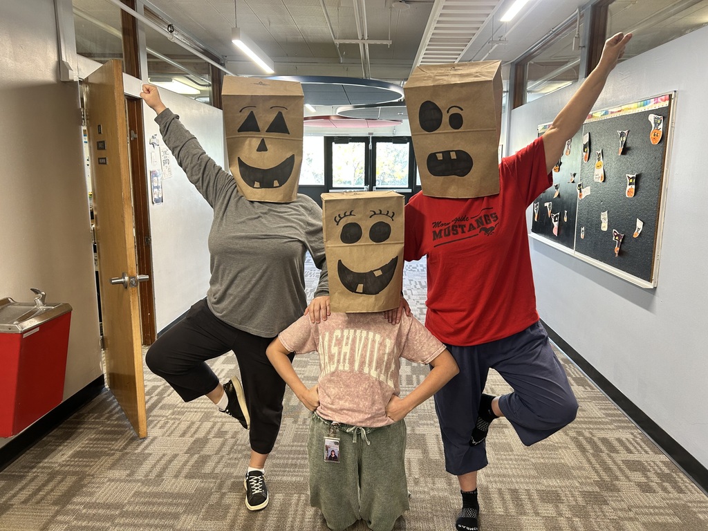 Three people wearing paper bag masks with faces on them pose in a hallway. One person crouches.