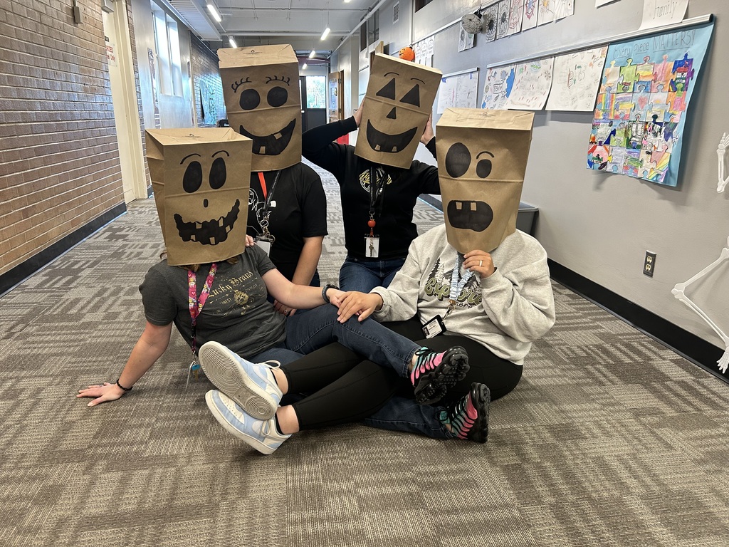 Four people sit on a floor wearing paper bags with drawn faces, one holding a bag.