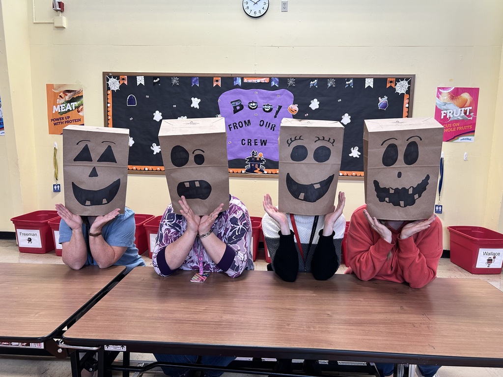 Four people sit at a table with paper bags over their heads; a blackboard with colorful posters is behind them.