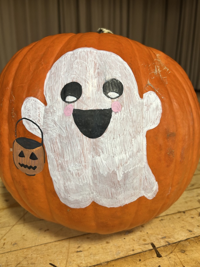 An orange pumpkin with a painted white ghost face sits on a wooden surface. The ghost face has eyes, a mouth, and a smile. A small pumpkin bucket is attached to the side.