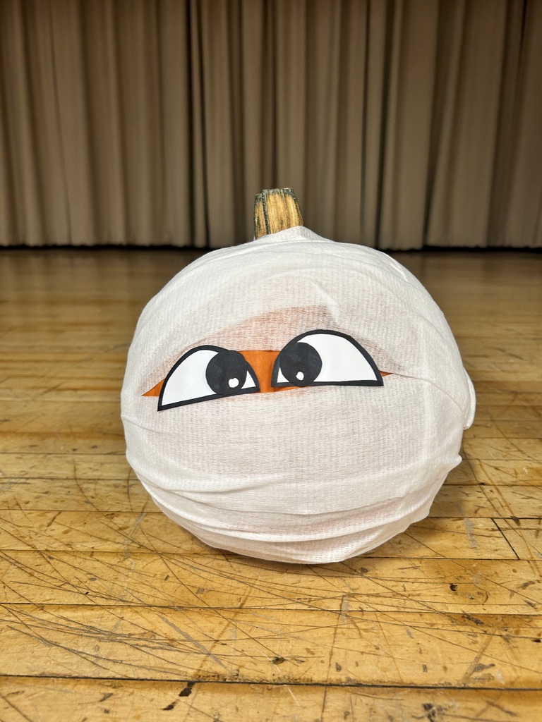 A pumpkin wrapped in white fabric with a face featuring large eyes and a small orange triangle on a wooden floor.