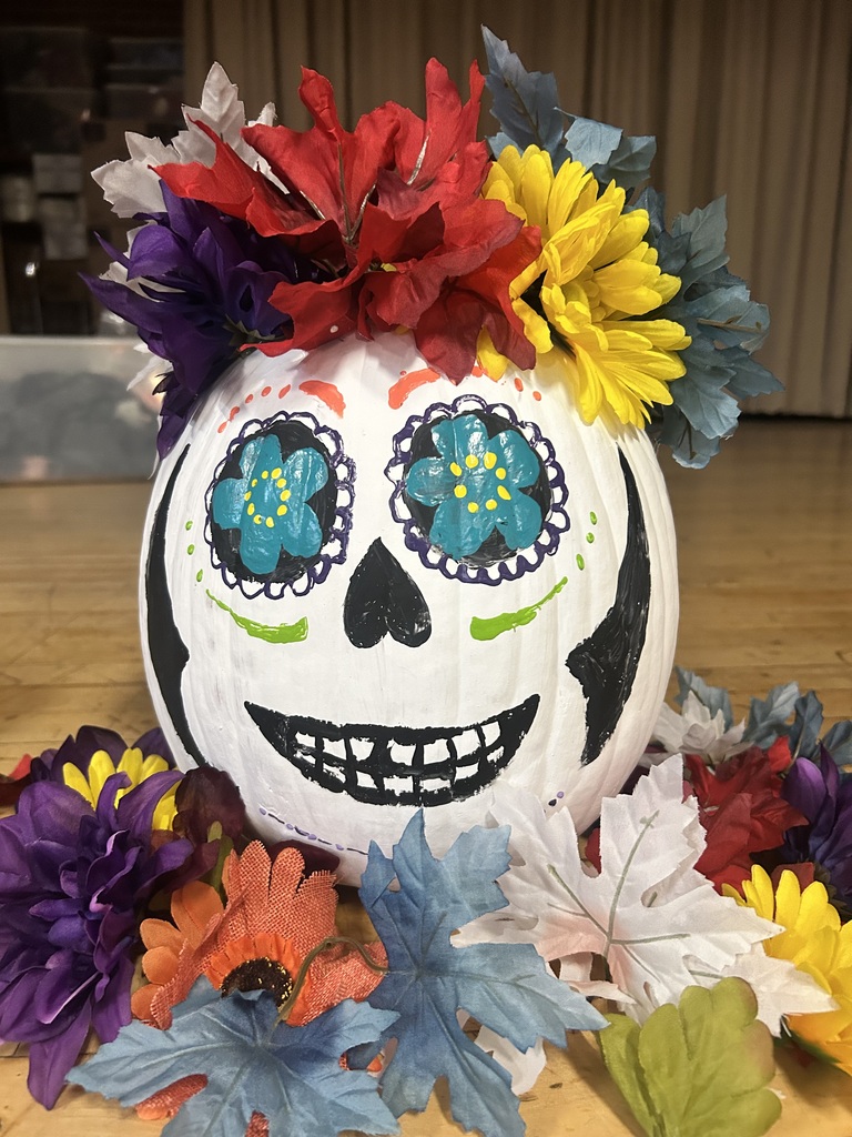 Painted pumpkin with skull design and colorful flowers on a table, surrounded by colorful paper flowers.