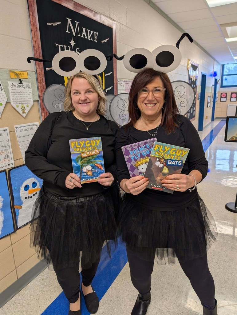 Two women dressed in matching fly-themed costumes hold "Fly Guy" children's books in a school hallway.