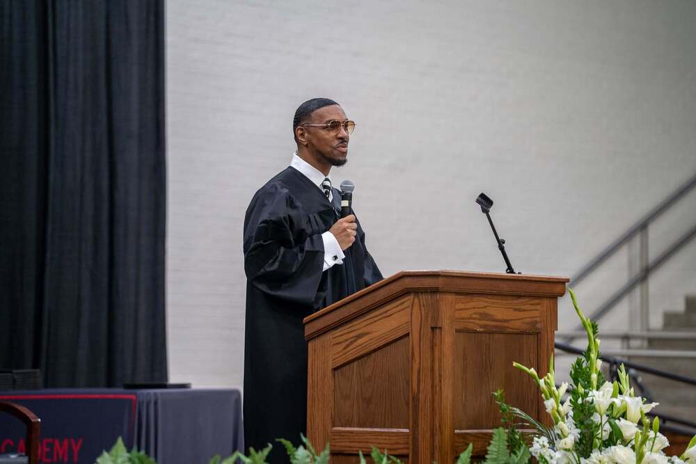 African American man wearing graduation gown standing behind a podium with a microphone in his hand